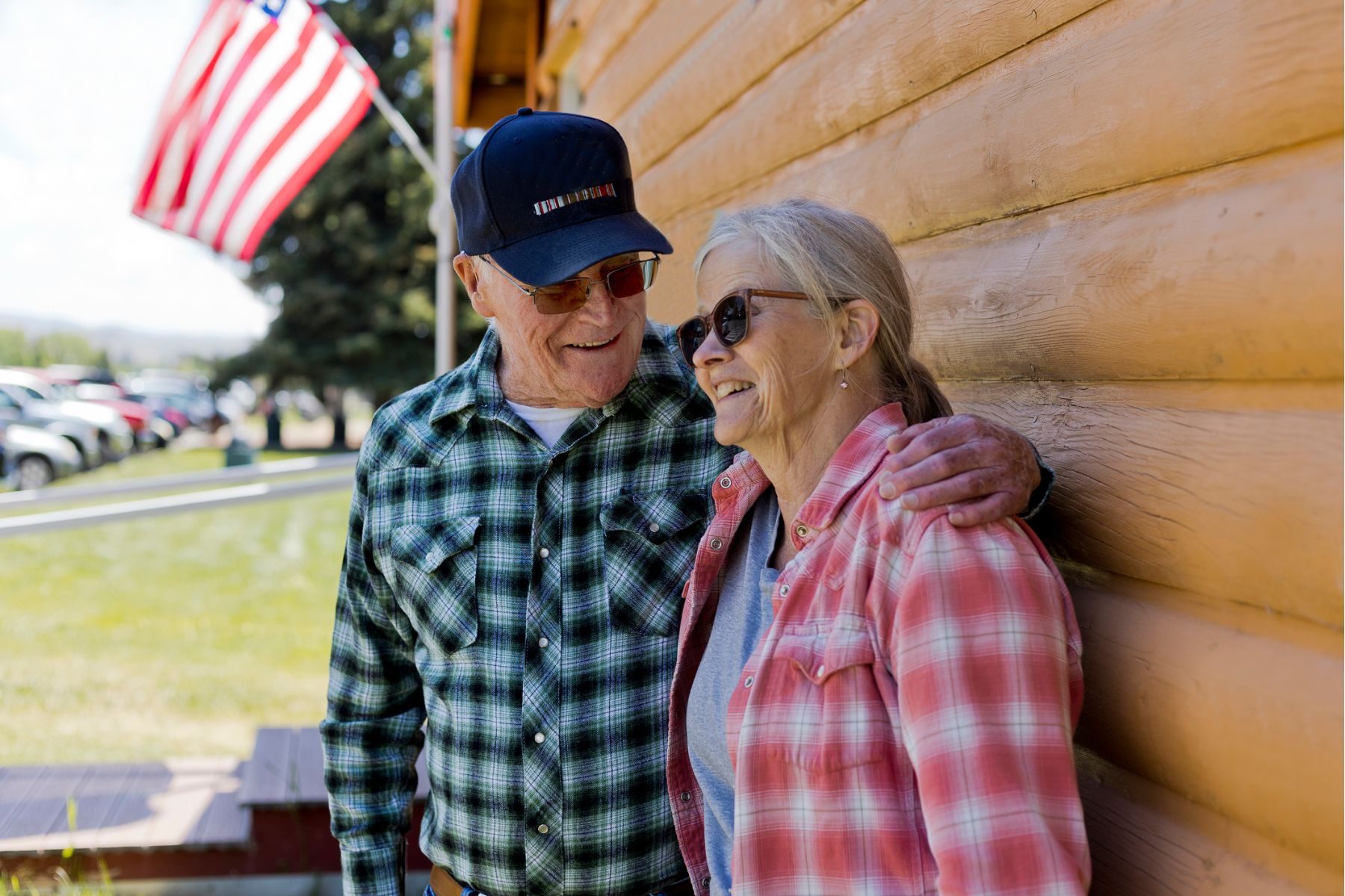 An older man and woman stand outside a wooden building, with a US flag in the background