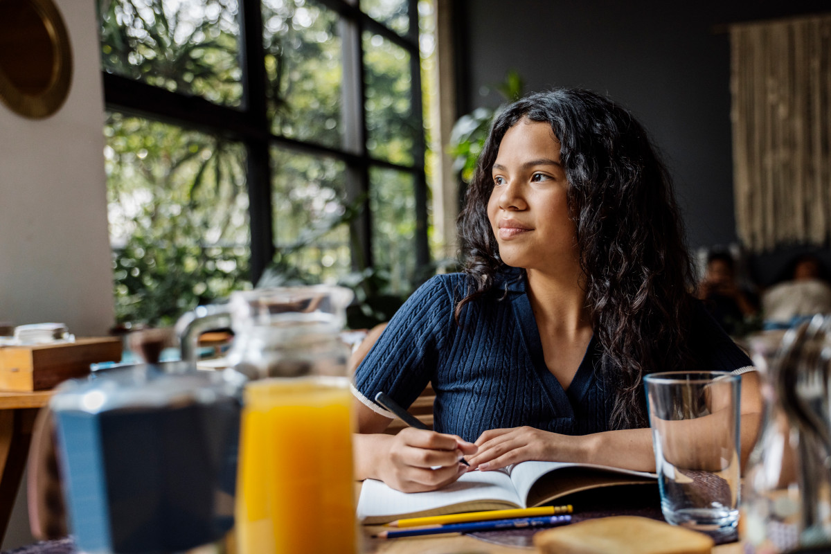 A young person sat up to a table, looking contemplatively out the window while writing in a journal/diary