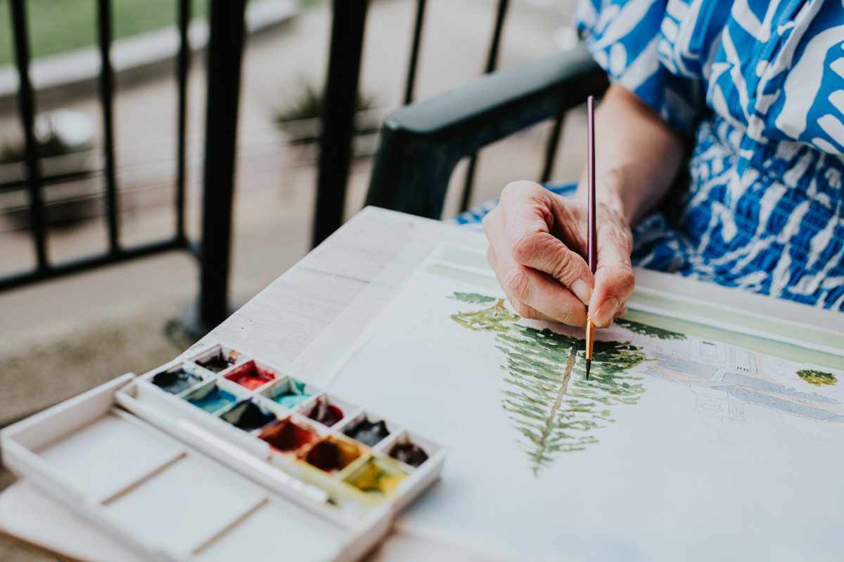 A woman dressed in a blue patterned dress engages in painting a serene tree landscape while seated on a balcony