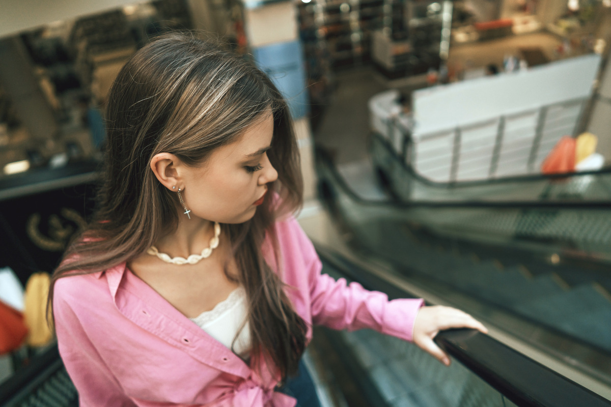 A young woman stands on an escalator in a bustling mall, wearing a pink shirt and shell necklace