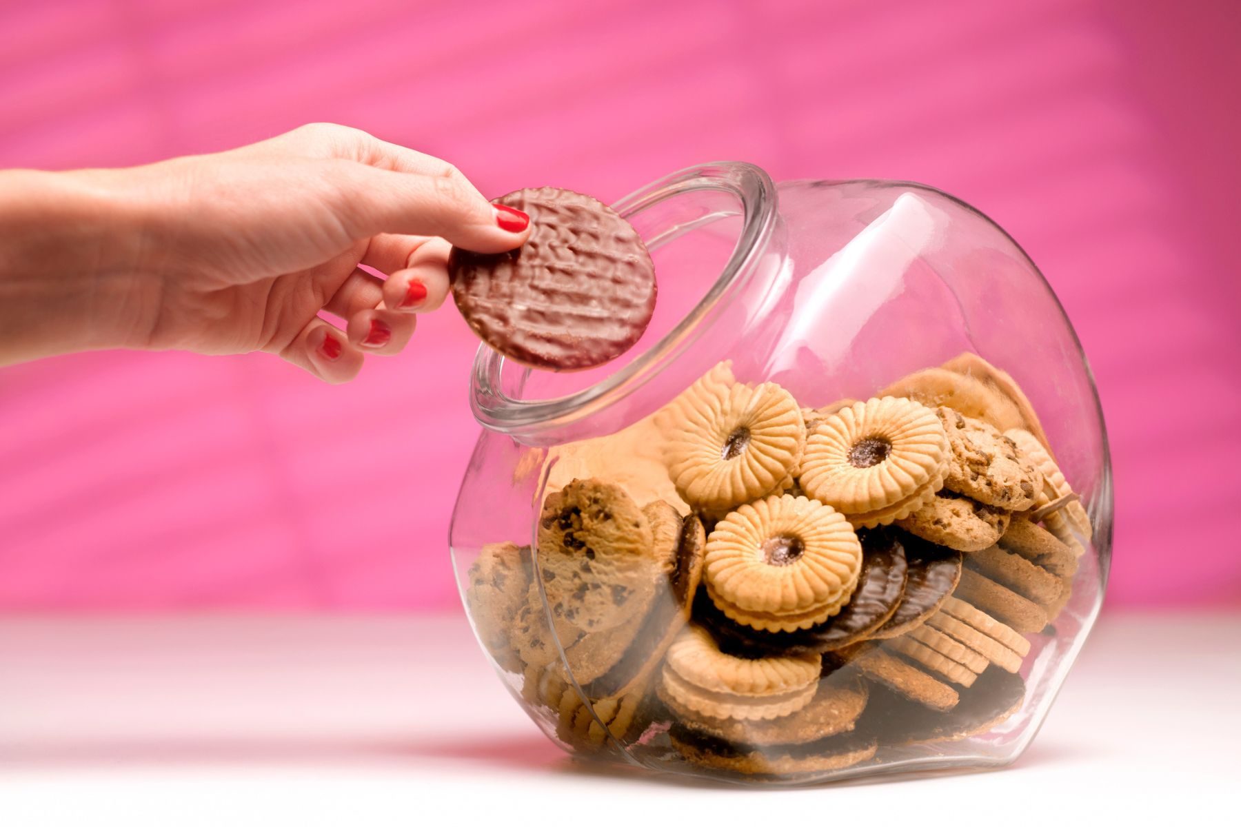 A hand takes a chocolate digestive biscuit out of a glass biscuit jar.