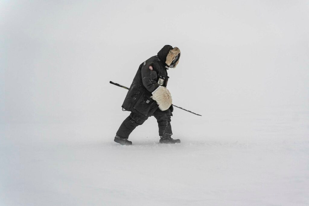 A side-on view of a person in extra-thick expedition gear walking with a drill in thick ice and snow