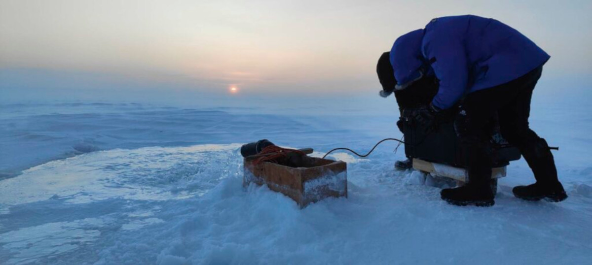 Two people wearing thick coats are hunched over and installing a battery in an icy and snowy landscape