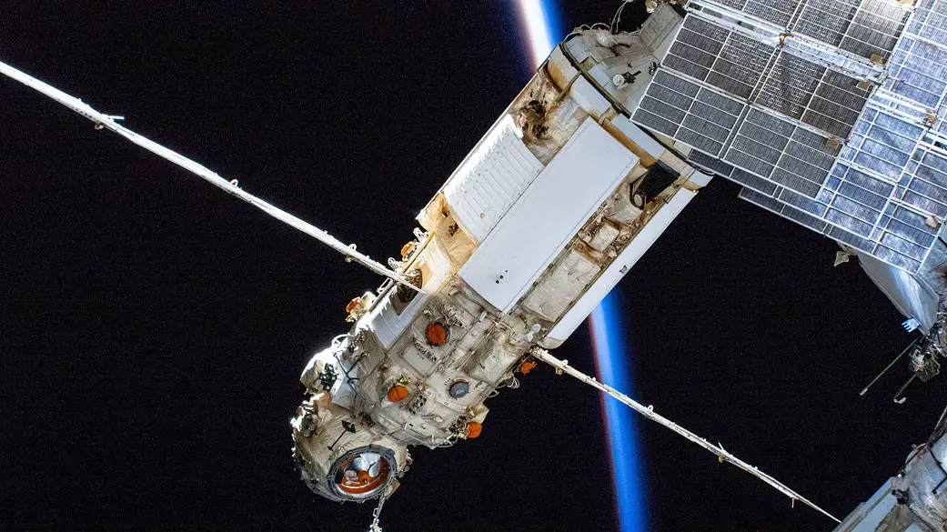 A white cylindrical module sticking out from the International Space Station. A backlit Earth is in the background.