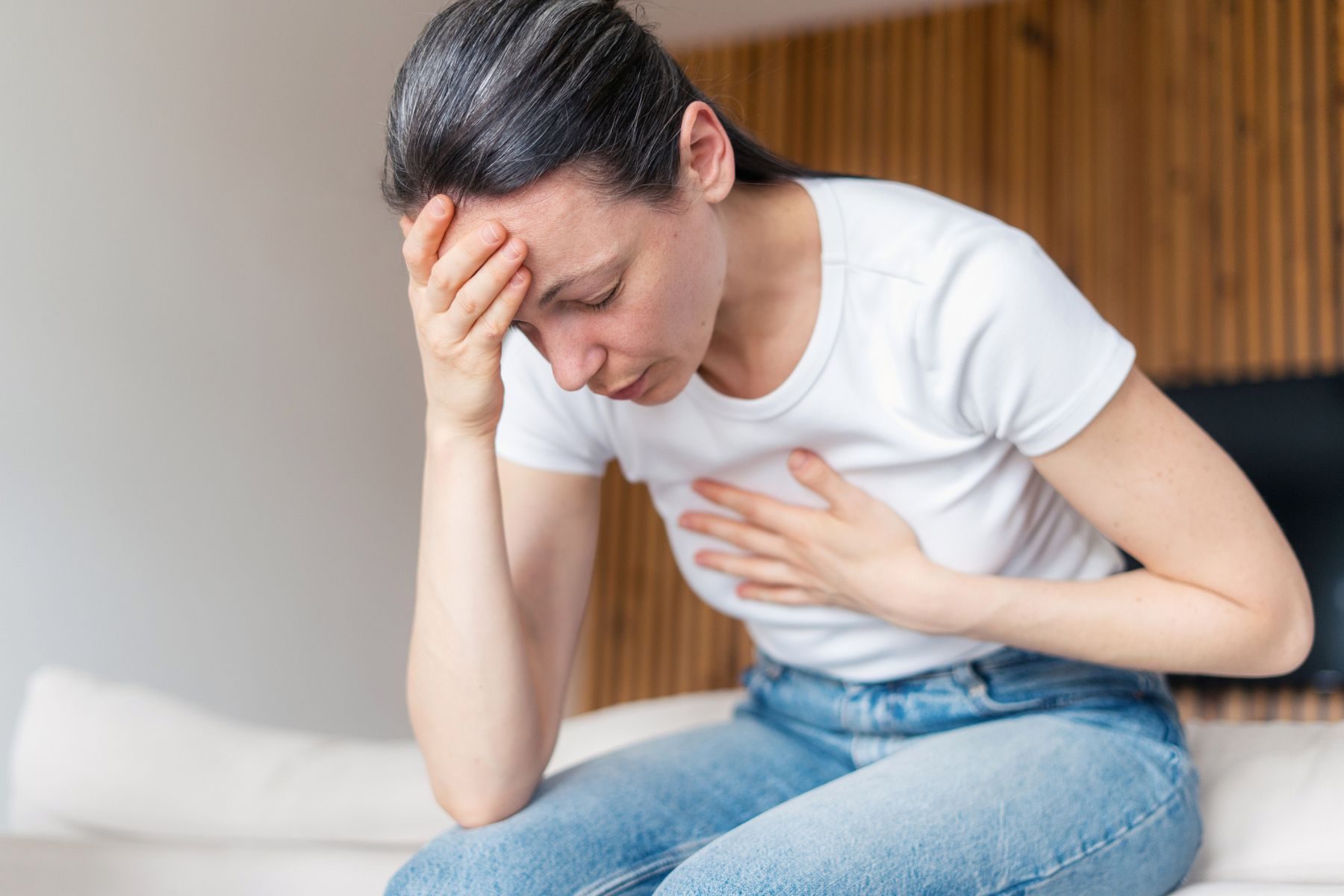 A woman feels unwell, sat on the side of a bed, holding her head and chest