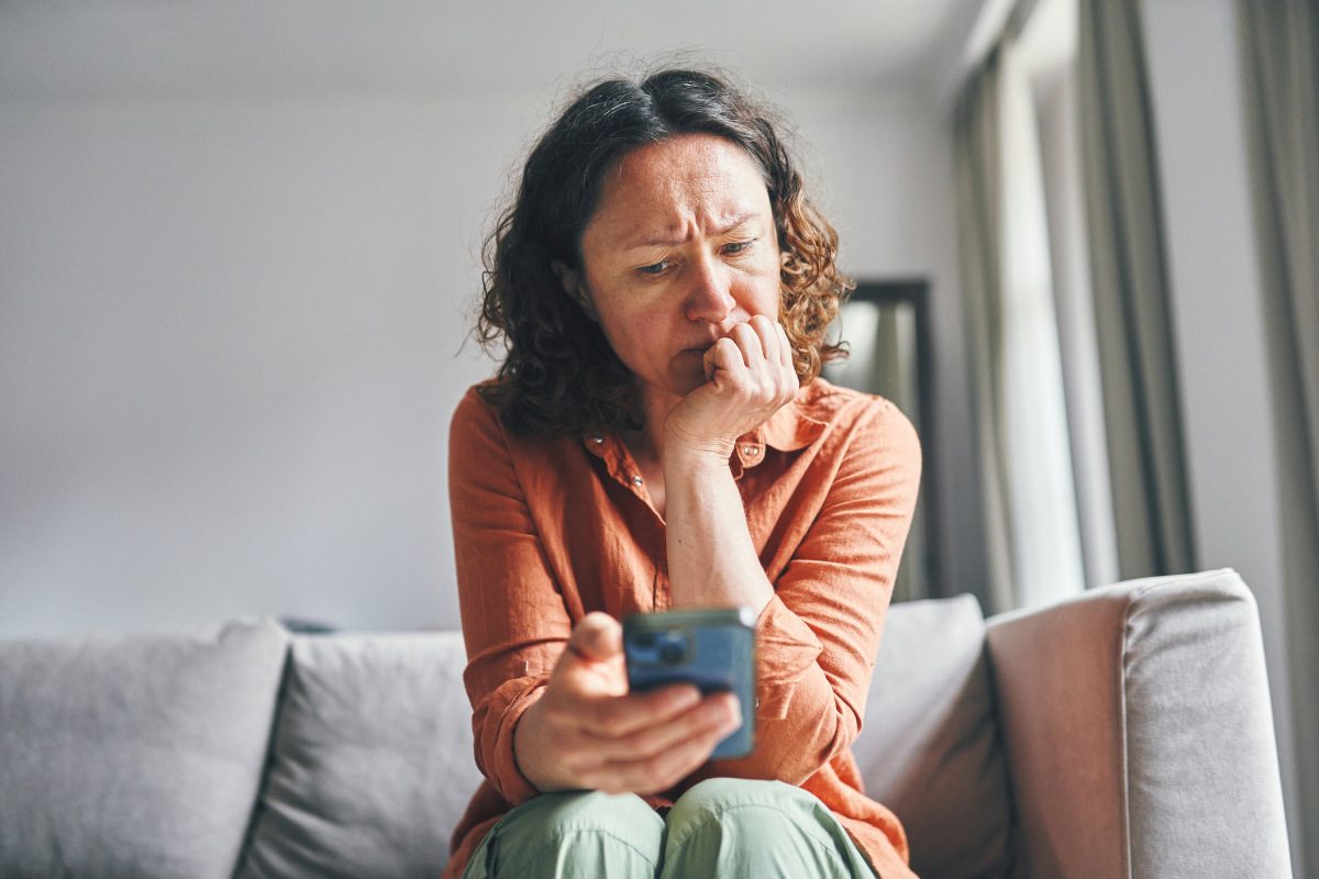 A woman sitting on a couch indoors, appearing focused and concerned while looking at her smartphone