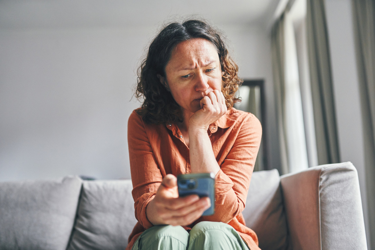 A woman sitting on a couch indoors, appearing focused and concerned while looking at her smartphone