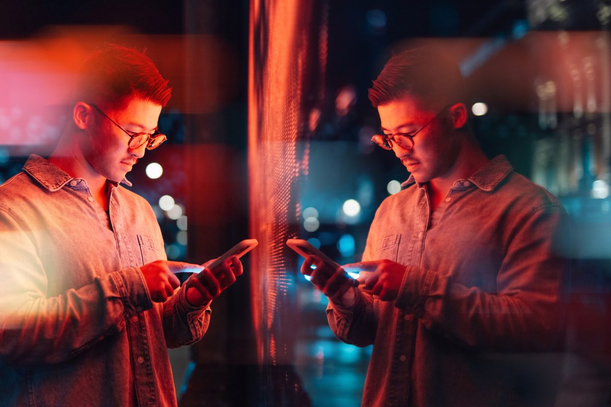 Side view of a young man using  smartphone, standing next to an illuminated red LED digital display screens in the city at night