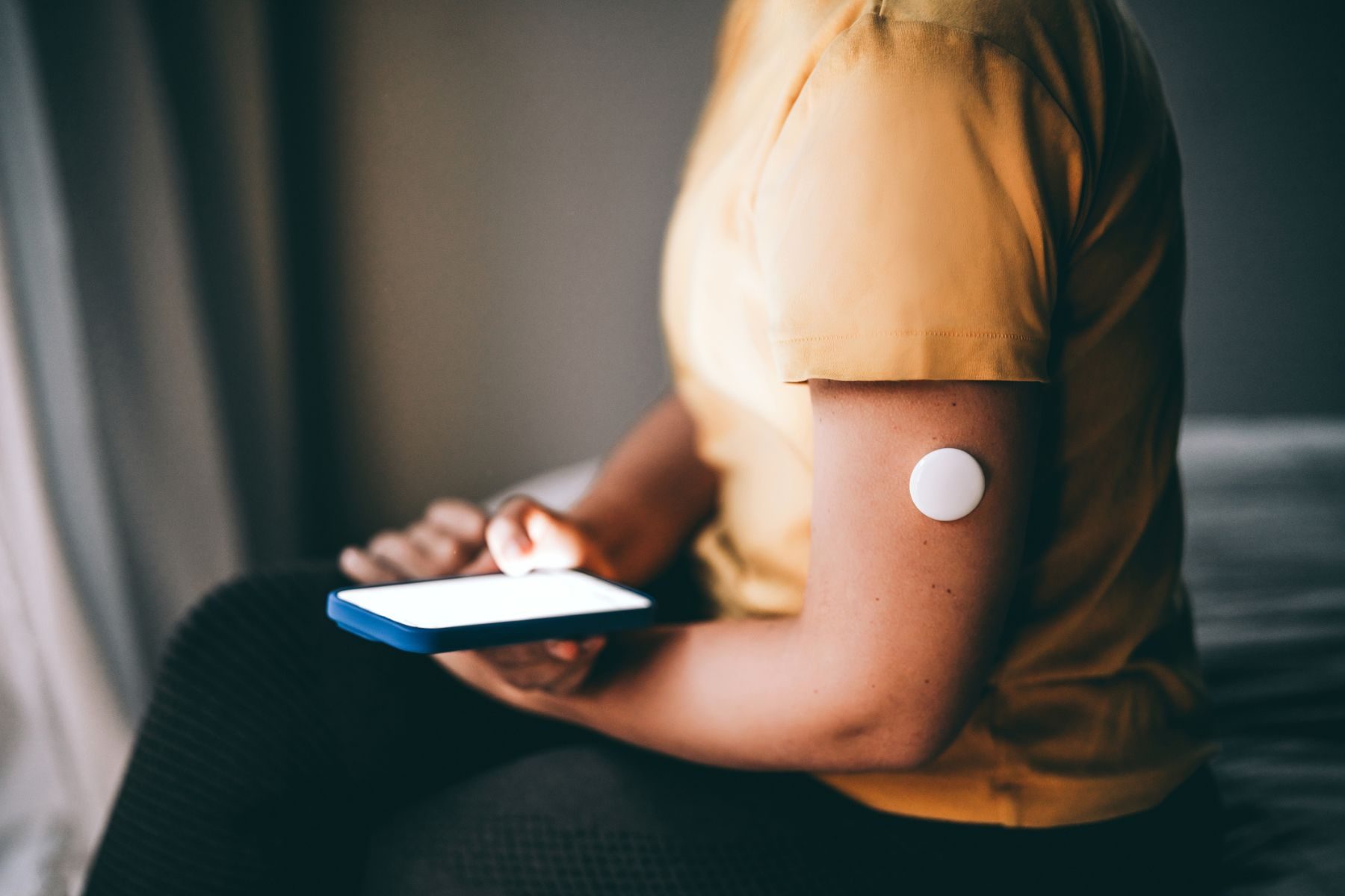 An unrecognisable person sits on a bed, with a continuous glucose monitor visible on their arm