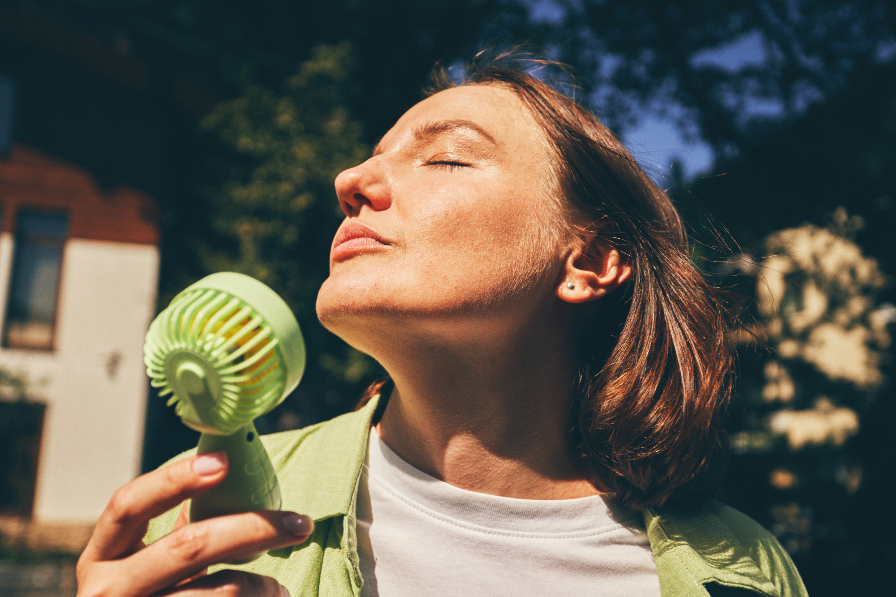 A woman holds a green portable fan to her face outdoors on a sunny day