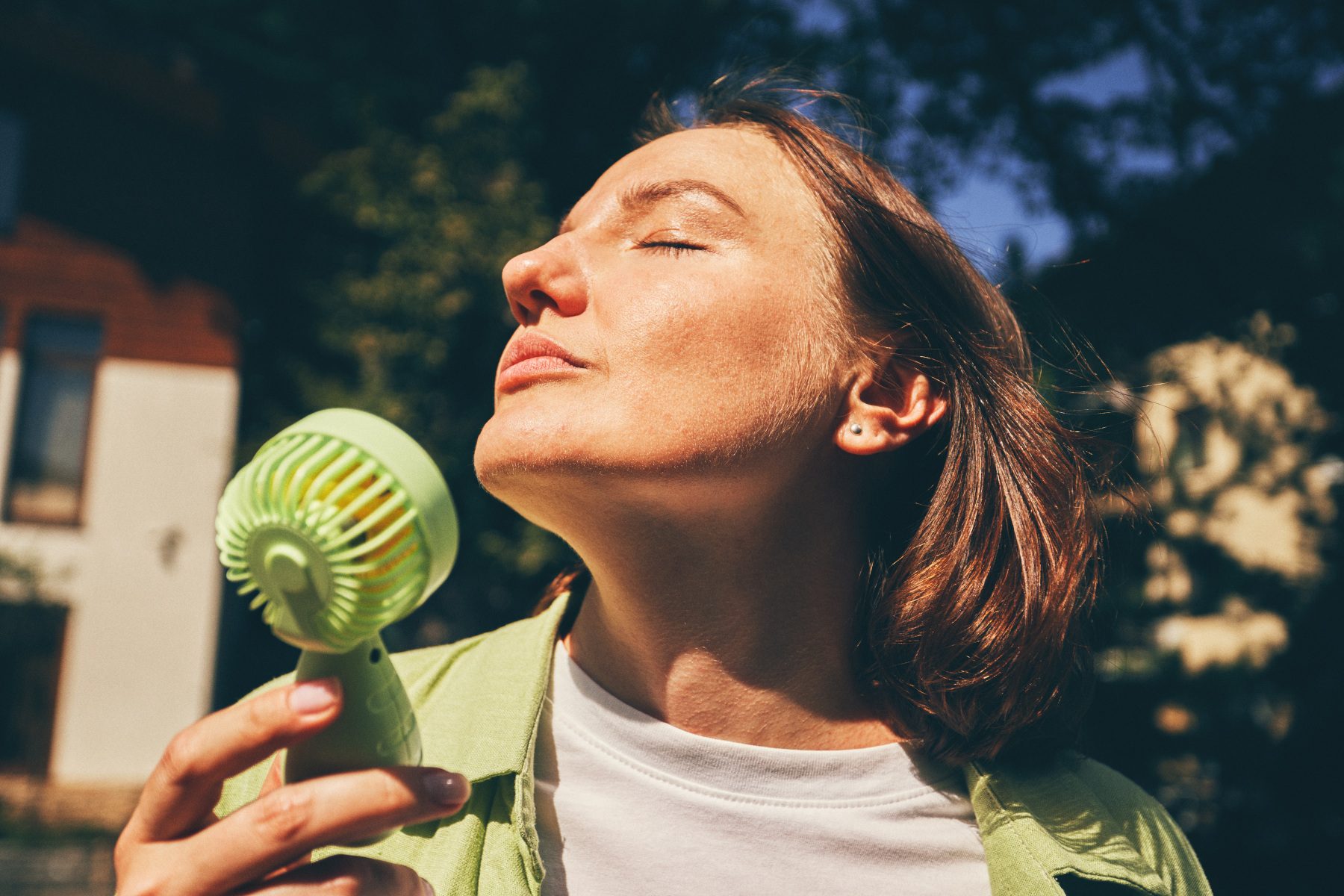 A woman holds a green portable fan to her face outdoors on a sunny day