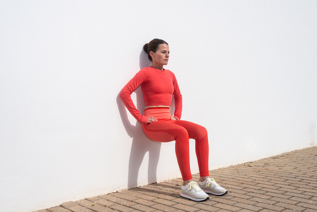 woman doing squats against a white wall outside in the sun.