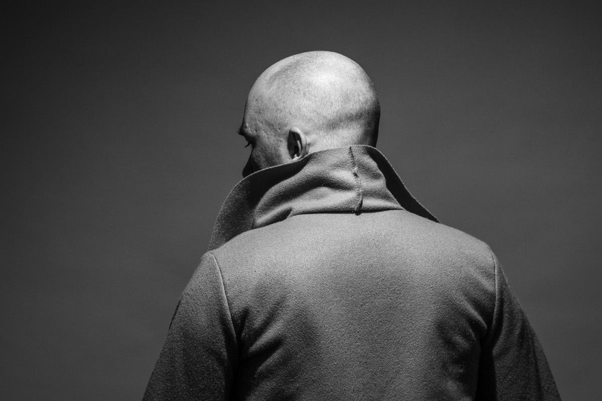 Black and white portrait of a mysterious bald man wearing a coat with the collar up, looking away from the camera against a dark background