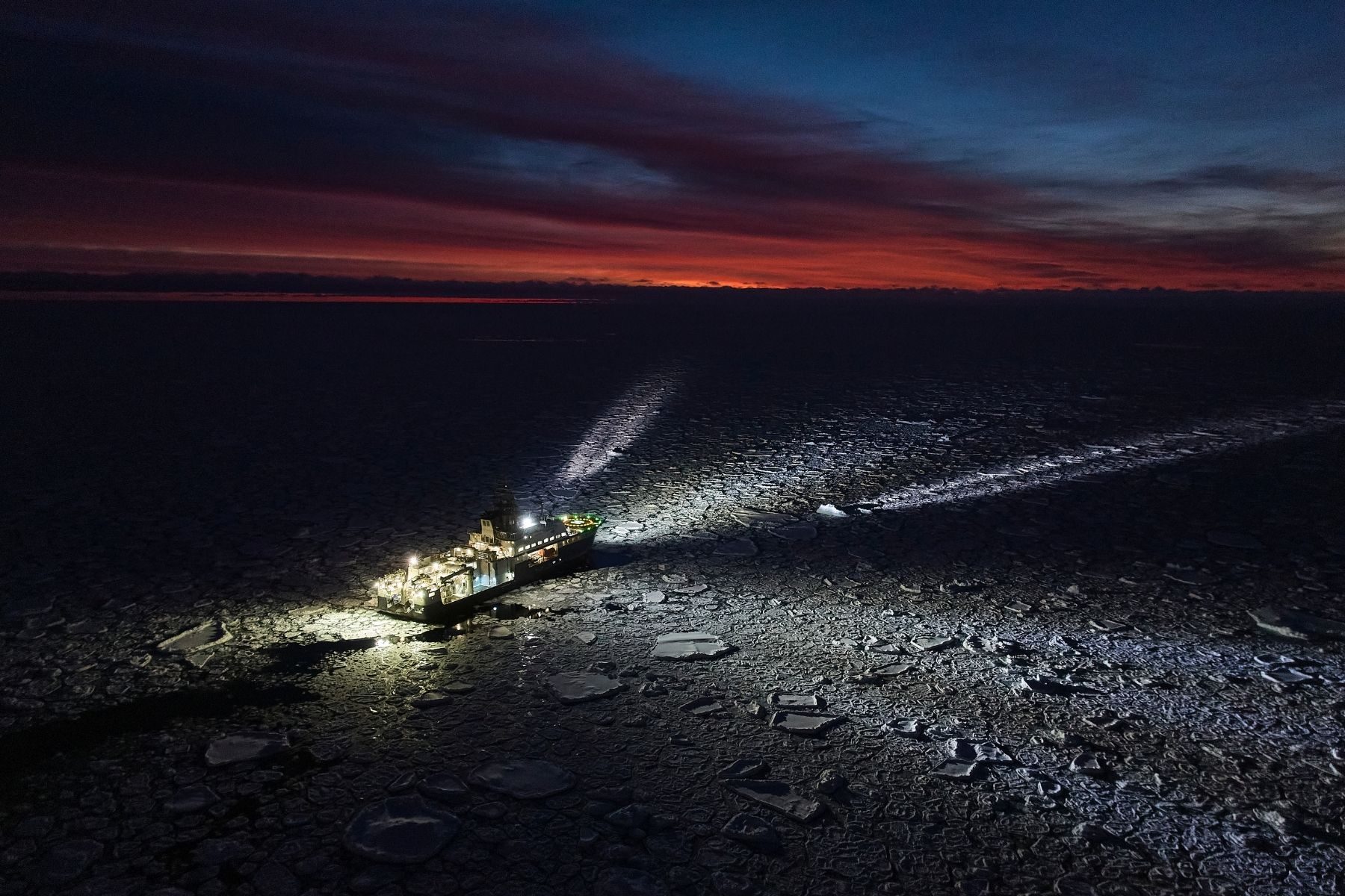 A large boat with its lights on sails through polar ice under a blue and red sky
