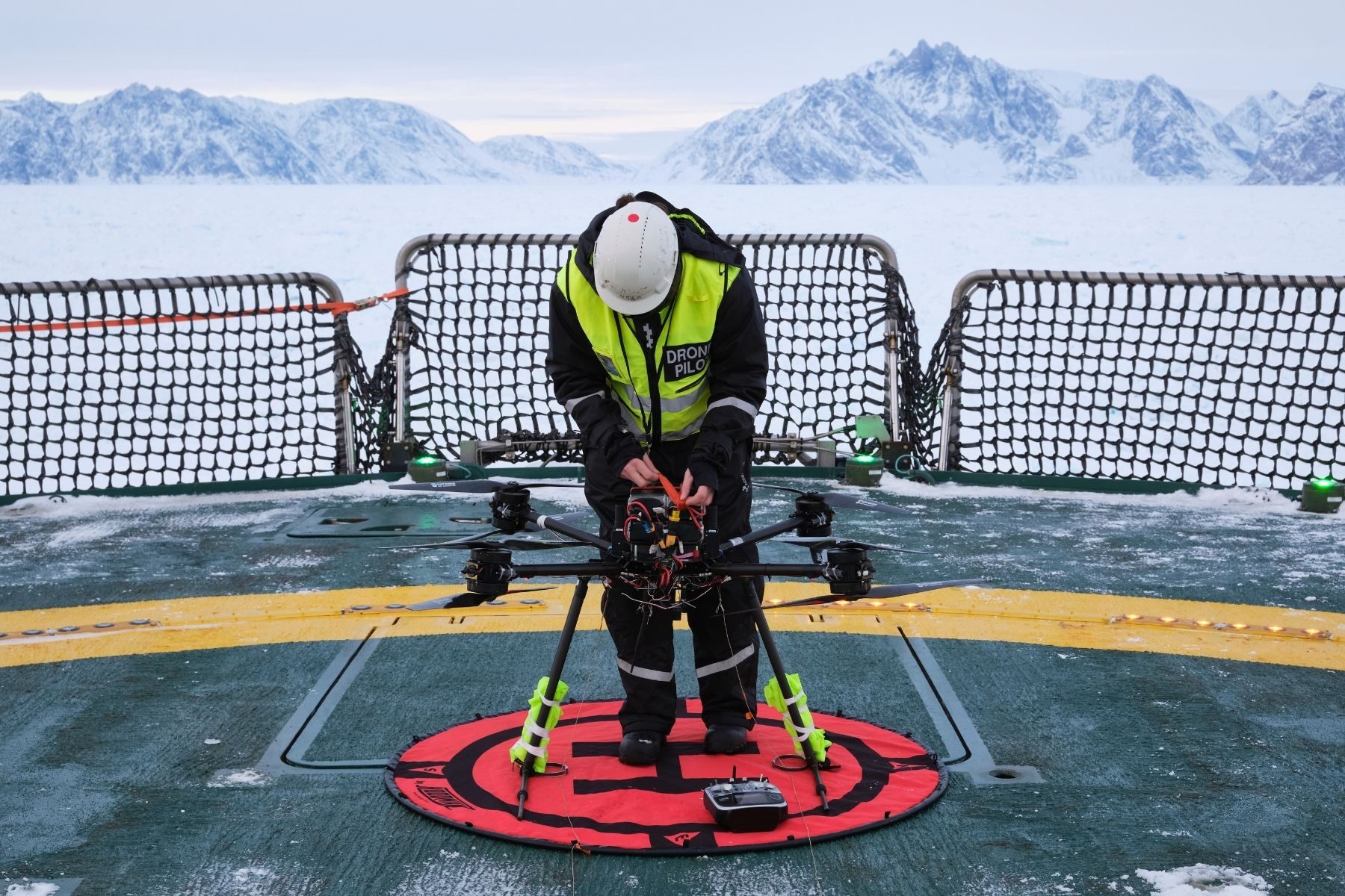 A person wearing a hard hat and a hi-vis vest sets up a drone on a ship, with icy flats and mountains in the background