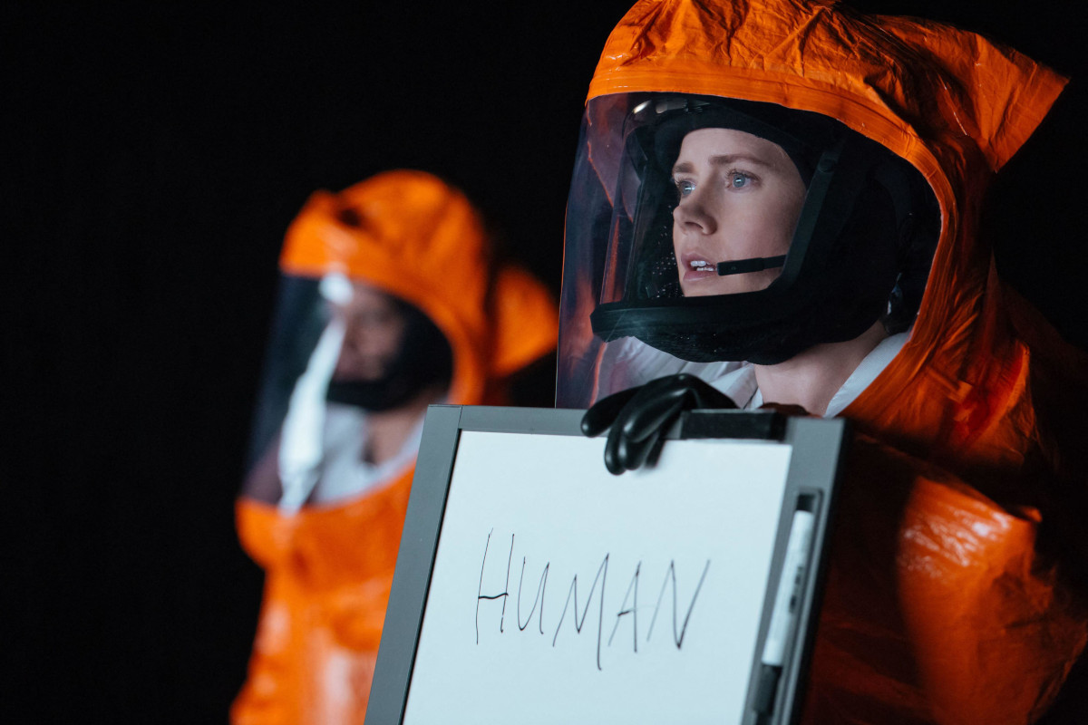 Screen from film 'Arrival', Amy Adams' character is in a hazmat suit and holding a whiteboard that she wrote 'human' on
