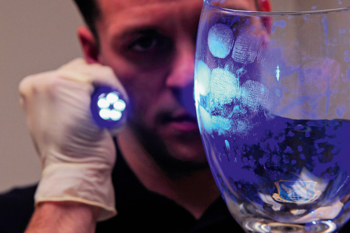 Forensics officer collecting evidence. Having dusted a glass with powder, the officer is using an ultraviolet (UV) torch to reveal the fingerprints.